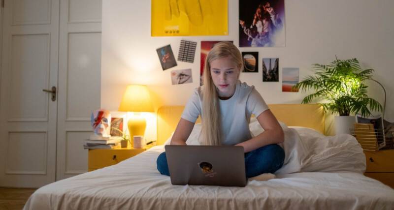 Nastolatka siedzi na łóżku w sypialni i korzysta z laptopa.