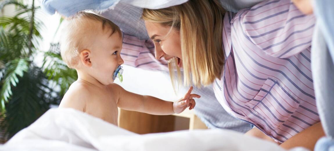 młoda kobieta w piżamie bawiąca się w chowanego z małym dzieckiem w łóżku