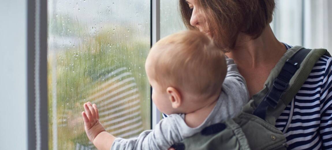 Młoda mama trzymająca niemowlę, patrząca w okno z wyrazem zmęczenia i refleksji, naturalne emocje po porodzie