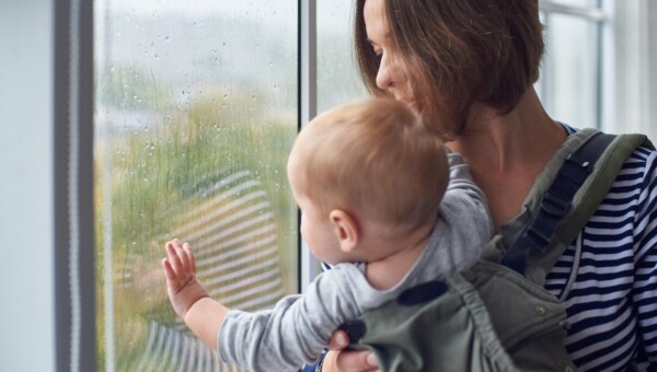 Młoda mama trzymająca niemowlę, patrząca w okno z wyrazem zmęczenia i refleksji, naturalne emocje po porodzie