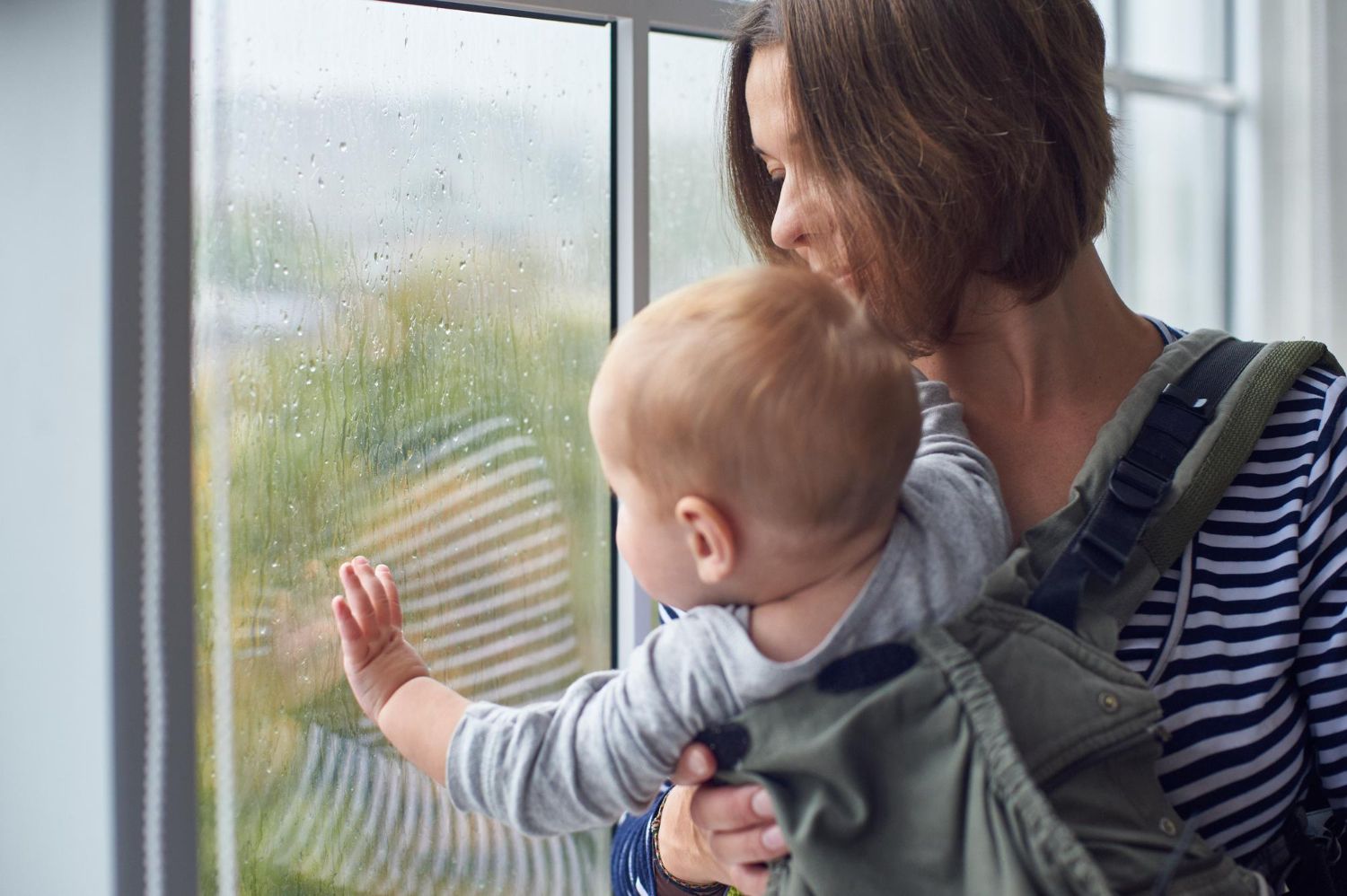 Młoda mama trzymająca niemowlę, patrząca w okno z wyrazem zmęczenia i refleksji, naturalne emocje po porodzie